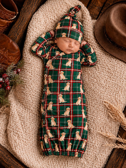 Sleeping baby in a green plaid outfit with puppy prints, resting on a cozy blanket with rustic decor around.