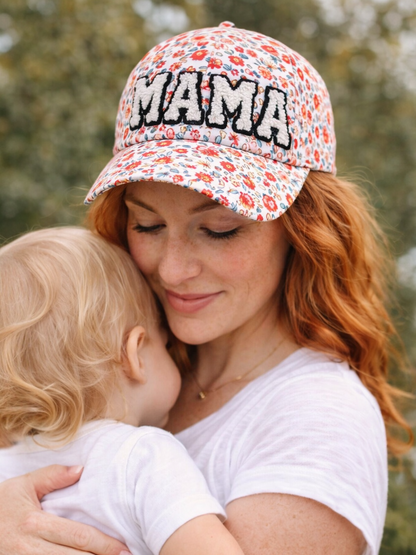 Mother cradles her child, wearing a floral cap with "MAMA" on it, in a soft, nature-filled background.