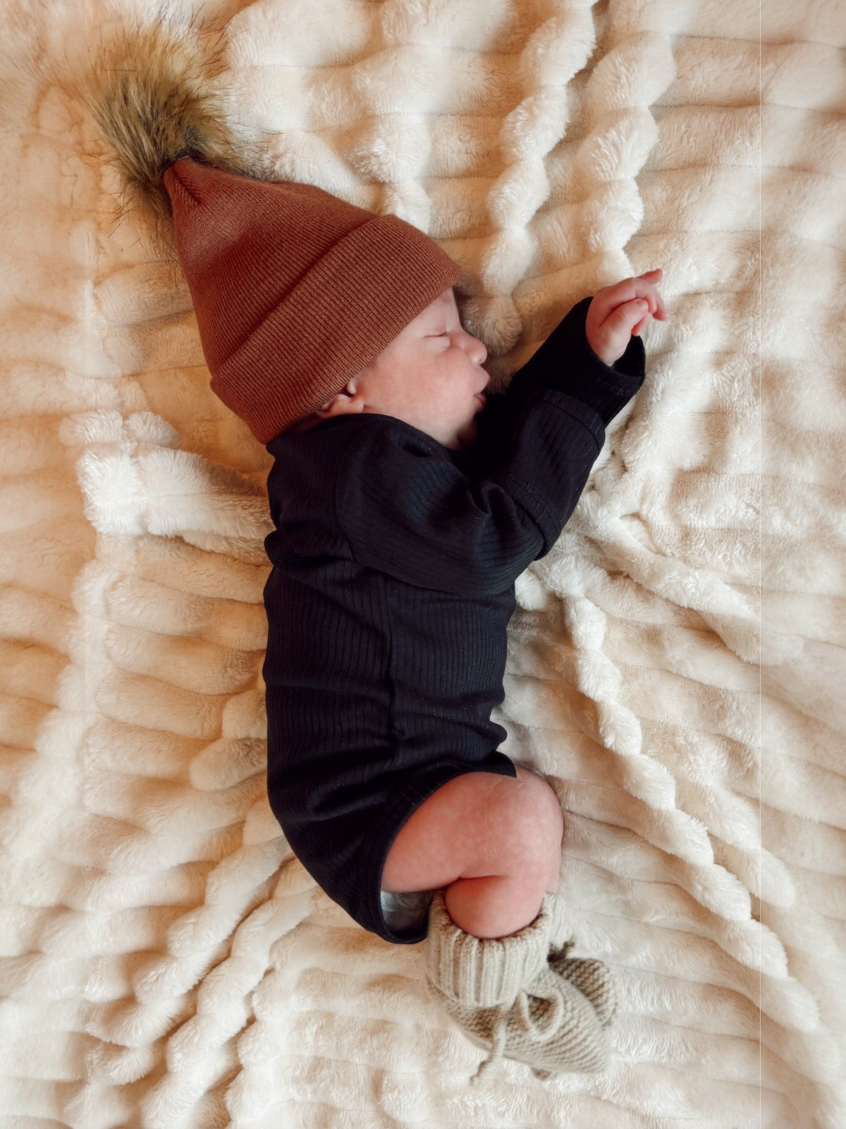 Newborn baby sleeping on a plush blanket, wearing a brown beanie and knitted booties, with a peaceful expression.