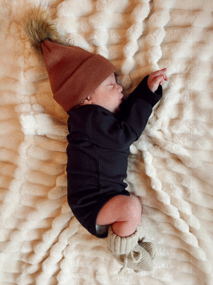 Newborn baby sleeping on a plush blanket, wearing a brown beanie and knitted booties, with a peaceful expression.