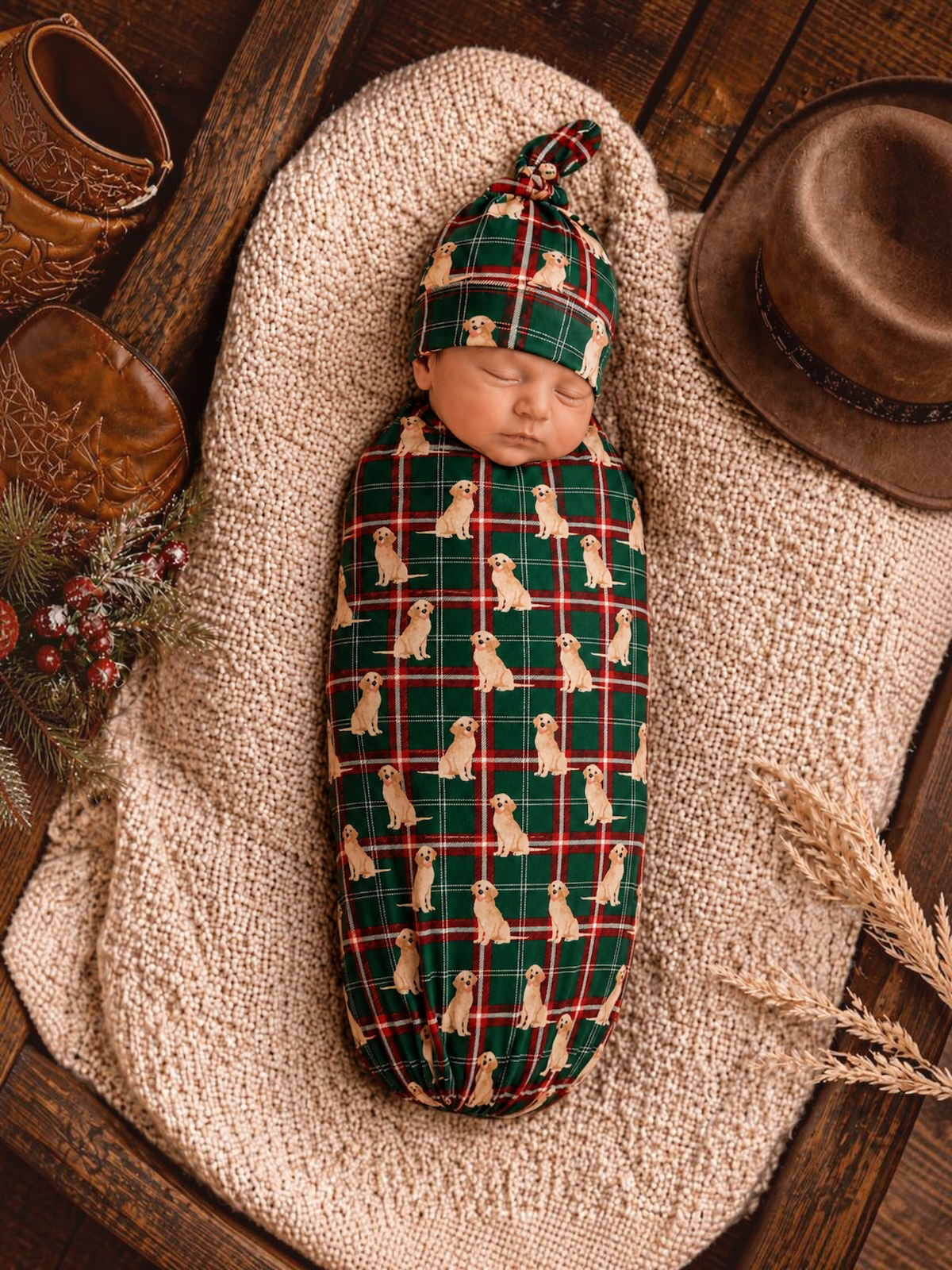 Newborn swaddled in a green plaid fabric with dog prints, resting on a textured blanket with rustic decor.