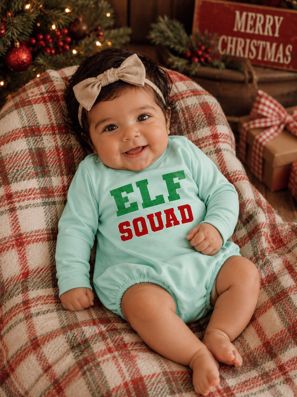 Smiling baby in mint outfit with "ELF SQUAD" text, sitting on plaid blanket near Christmas decor.