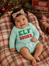 Smiling baby in mint outfit with "ELF SQUAD" text, sitting on plaid blanket near Christmas decor.