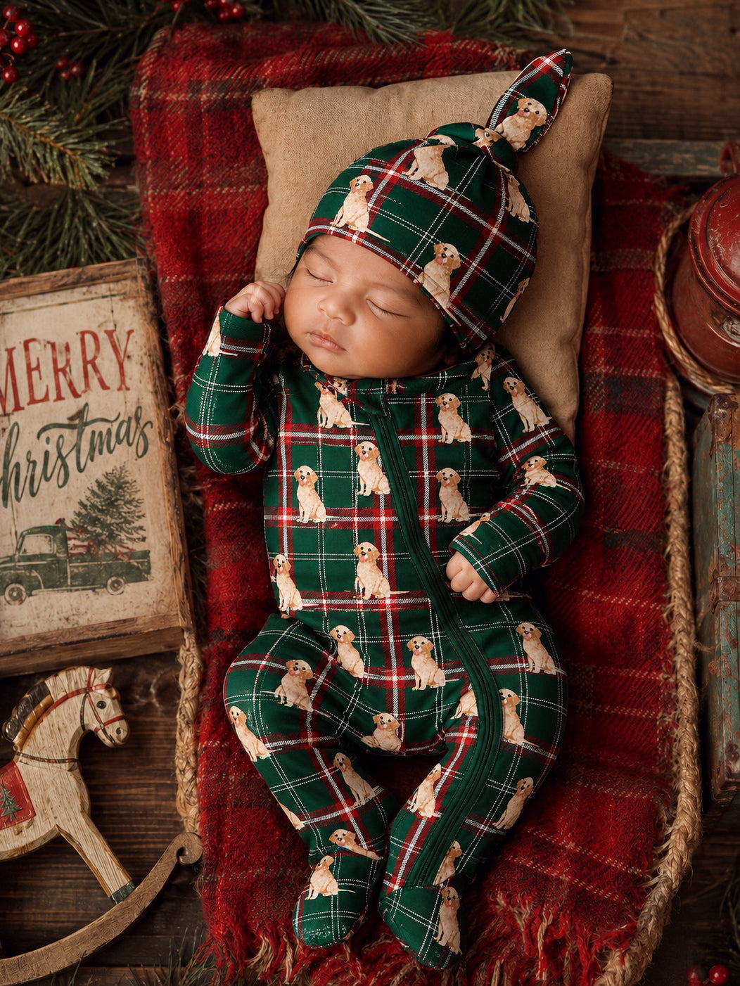 Baby sleeping on a cozy blanket, wearing a green plaid outfit with puppy prints and a matching hat, festive decor in background.