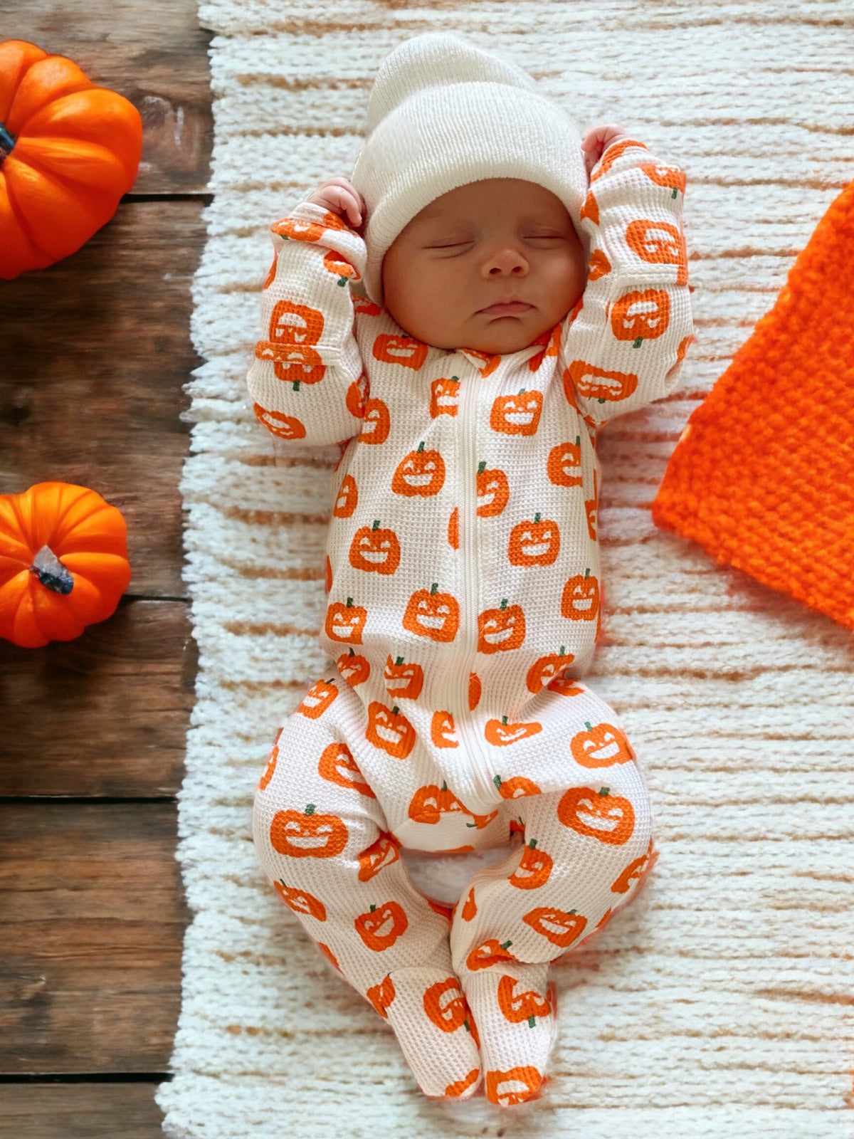 Newborn in pumpkin-patterned outfit, sleeping on a textured blanket, surrounded by small pumpkins and an orange cloth.