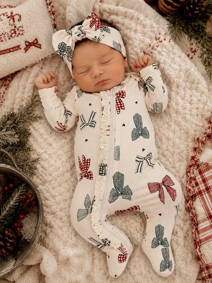 Sleeping baby in a festive onesie with bows, surrounded by cozy blankets and holiday decorations.