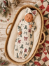 Newborn sleeping in a cozy basket, wearing a festive onesie and bow, surrounded by holiday decor.