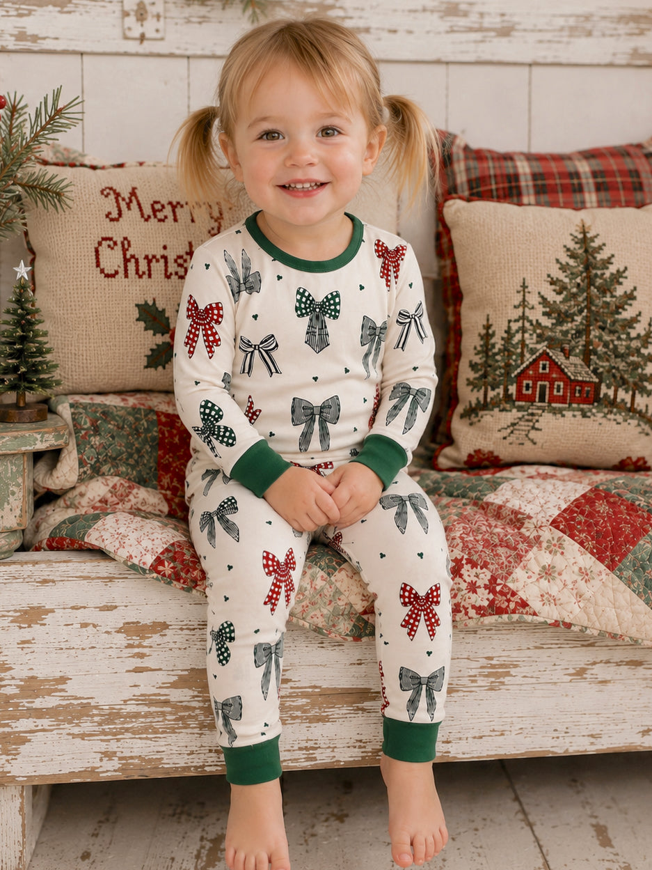 Smiling toddler in festive pajamas with bows, sitting on a quilted blanket among Christmas decorations.