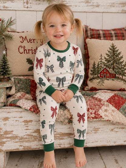 Smiling toddler in festive pajamas with bows, sitting on a quilted blanket among Christmas decorations.