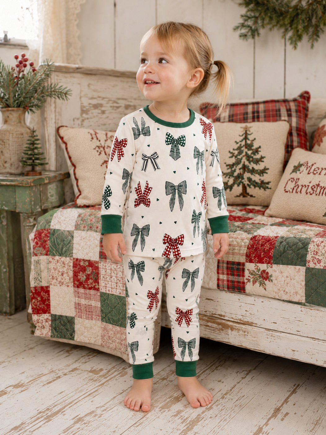 Smiling child in festive pajamas with bows, standing on a wooden floor near a quilted couch.
