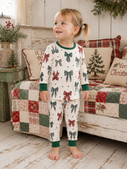 Smiling child in festive pajamas with bows, standing on a wooden floor near a quilted couch.
