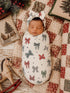 Infant swaddled in a bow-patterned blanket, resting on a quilt with holiday decor and a wooden rocking horse.