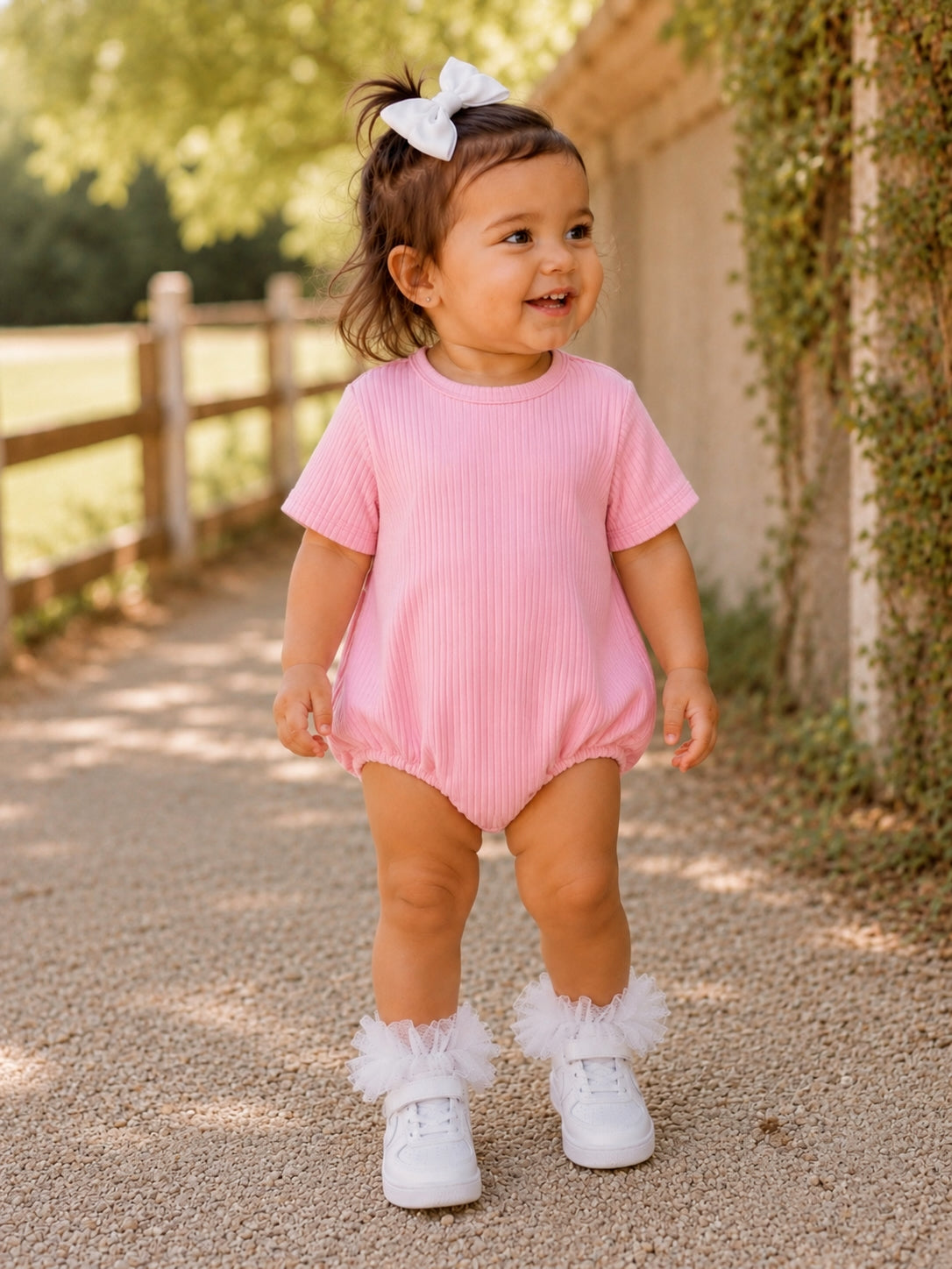Smiling toddler in a pink outfit and fluffy white shoes stands on a gravel path, surrounded by greenery.