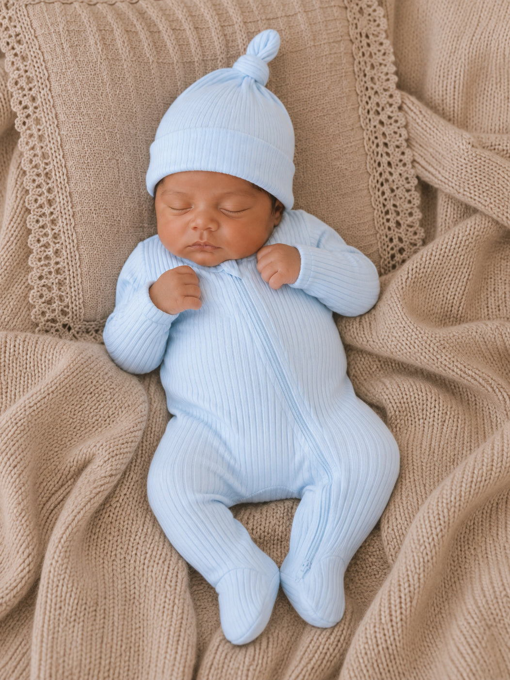 Sleeping baby in a light blue outfit and hat, nestled on a cozy beige blanket.