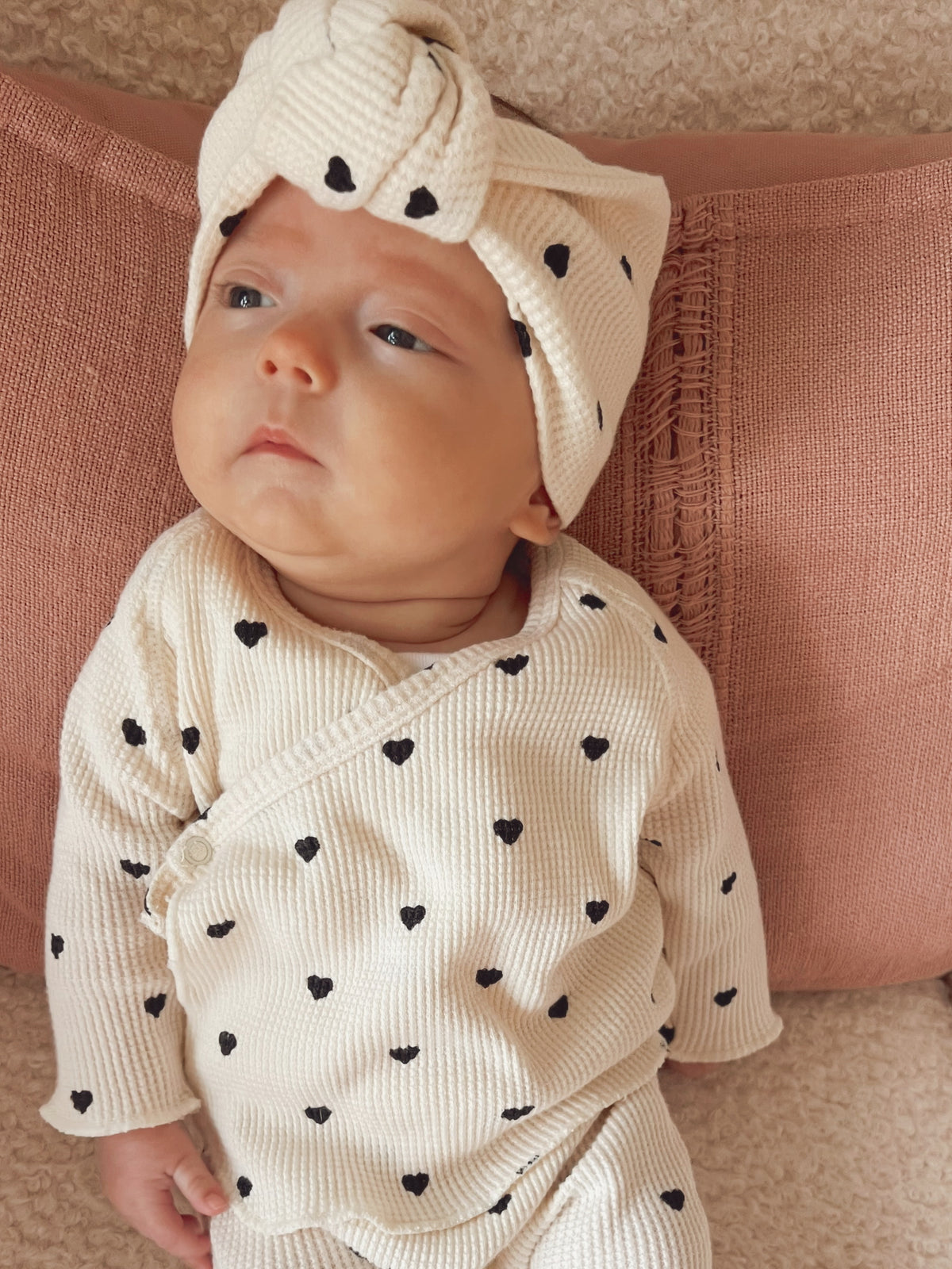 Baby with a headband and heart-patterned outfit lying on a pink pillow, looking curiously at the camera.