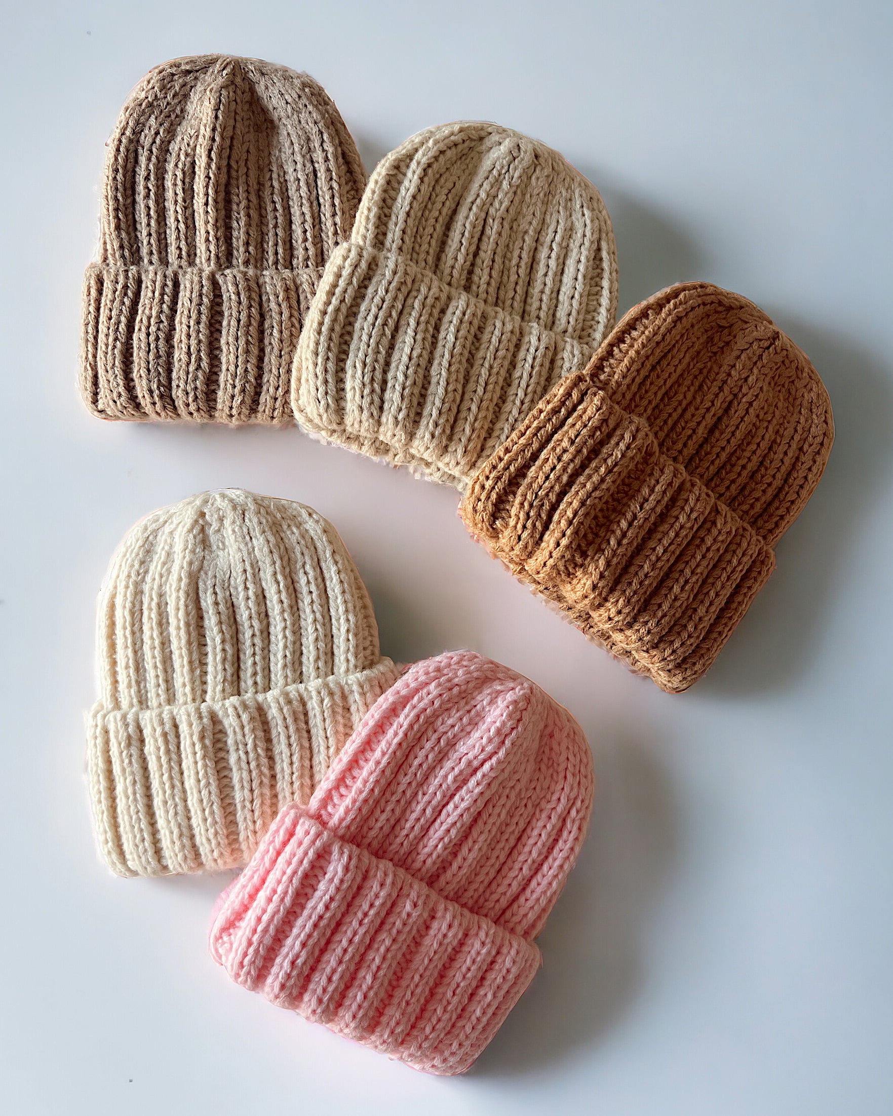 Five knitted beanies in cream, pink, light brown, and dark brown arranged on a white surface.