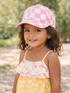 Smiling child with curly hair wearing a pink and white checkered cap and floral summer dress in a natural setting.