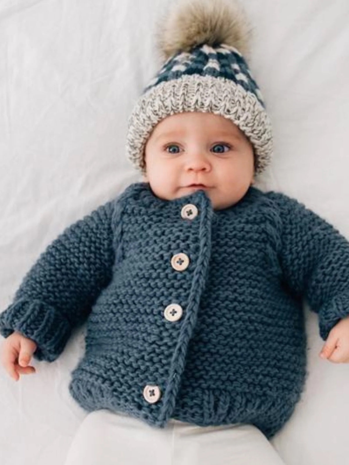 Baby wearing a cozy knitted gray jacket and a patterned hat with a pom-pom, resting on a white background.