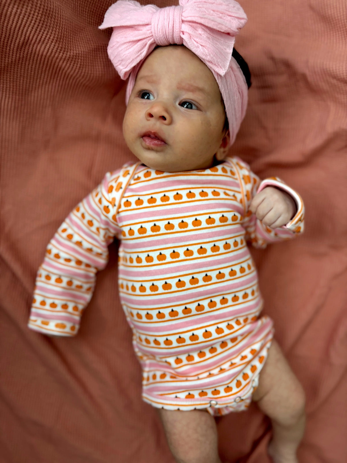 Baby in pumpkin-patterned onesie and large pink bow on a soft pink background.