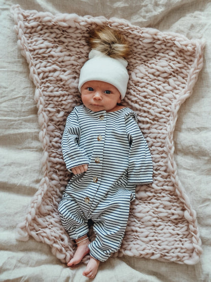 Infant in striped onesie and white hat, lying on a pink knitted blanket.