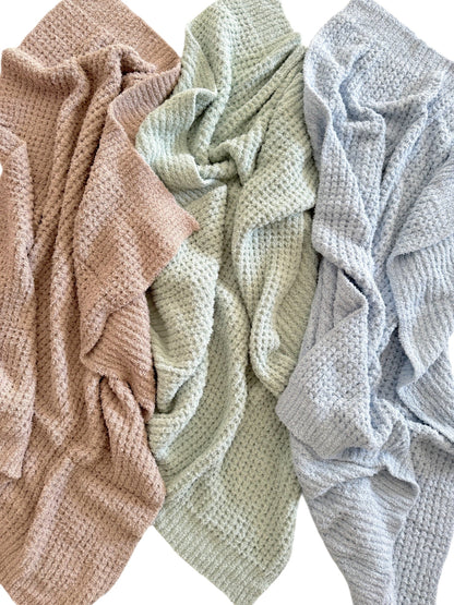 Three textured blankets in soft pastel colors: pink, mint green, and light blue, arranged casually.