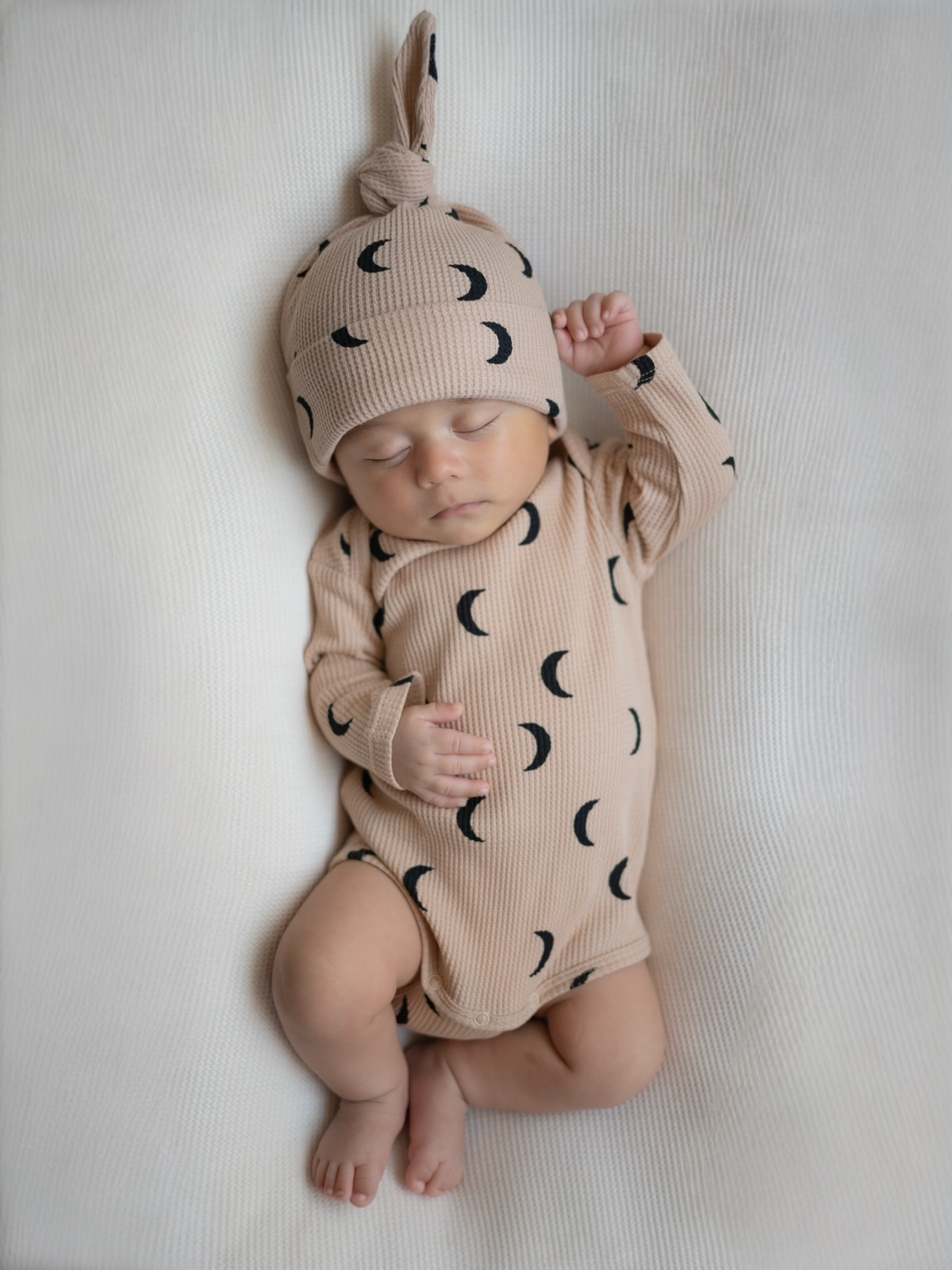 Sleeping baby in a crescent moon patterned onesie and hat on a textured white blanket.