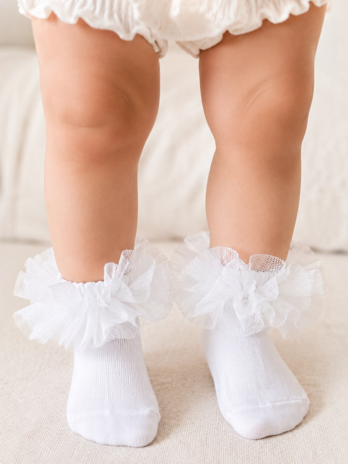 Baby legs in white socks with tulle ruffles, resting on a soft surface.