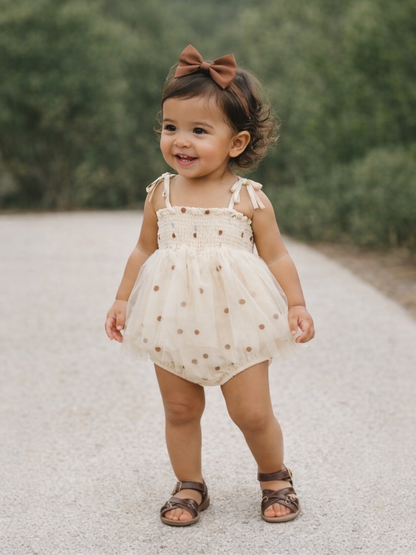 Smiling toddler in a polka dot outfit and a bow, standing on a gravel path surrounded by greenery.