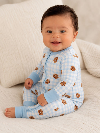 Smiling baby in blue checked pajamas with teddy bear prints sitting on a soft blanket.