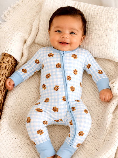 Smiling baby in cute blue checkered onesie with teddy bear print, lying on a cozy blanket.