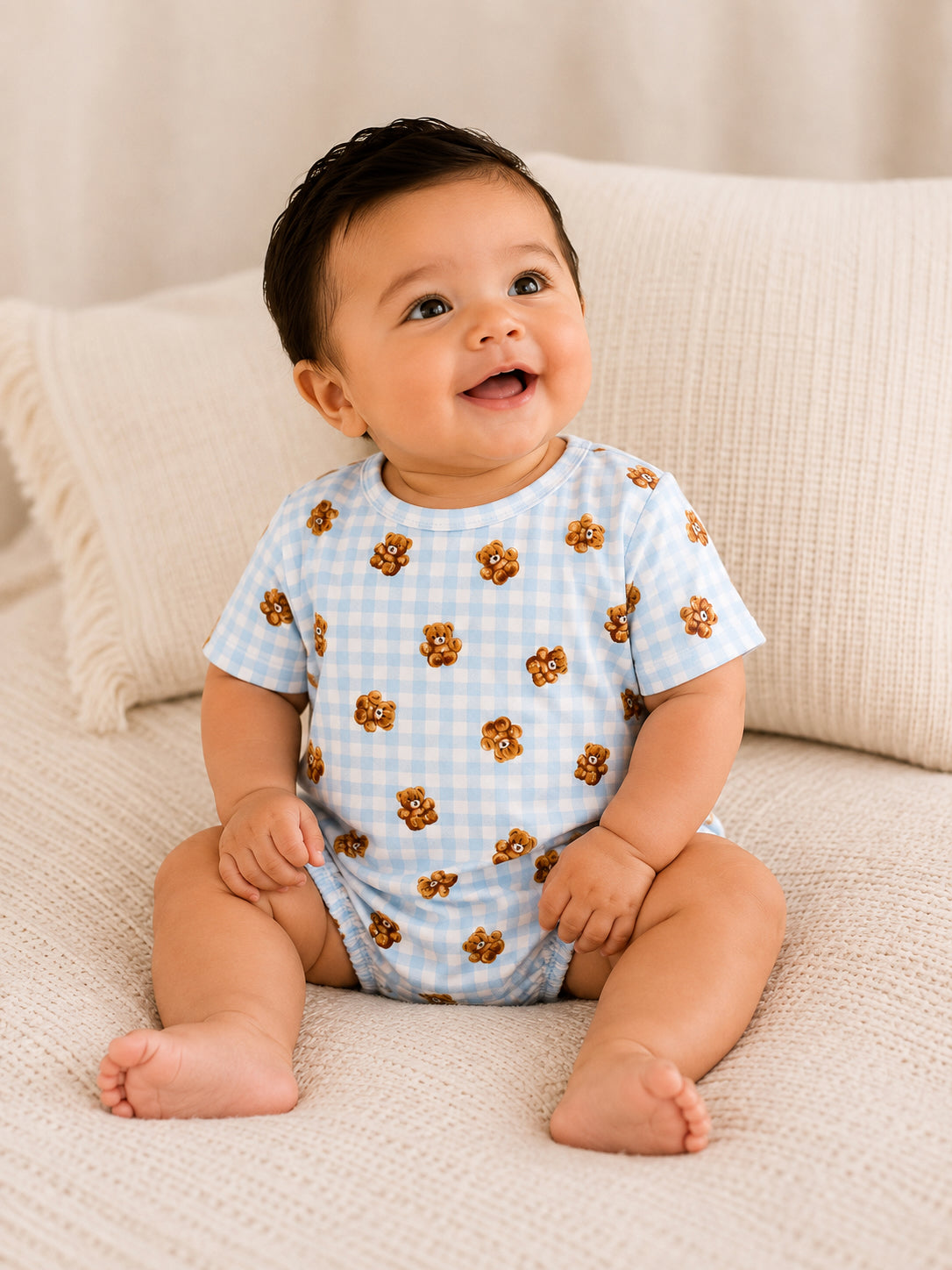 Smiling baby in a blue checked onesie with bear print, seated on a cozy beige blanket.