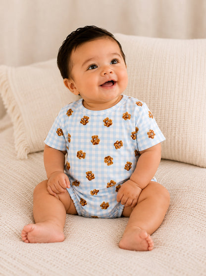 Smiling baby in a blue checked onesie with bear print, seated on a cozy beige blanket.