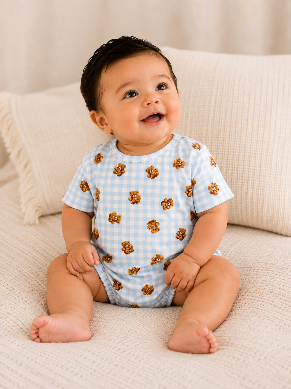 Smiling baby in a blue checked onesie with bear print, seated on a cozy beige blanket.