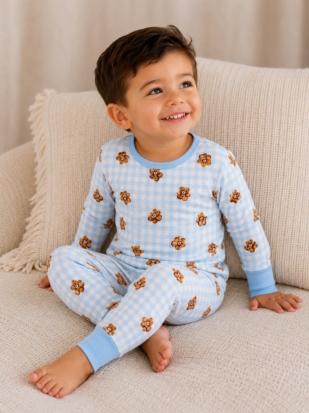 Smiling young boy in blue checkered pajamas with lion patterns, sitting on a cozy beige sofa.