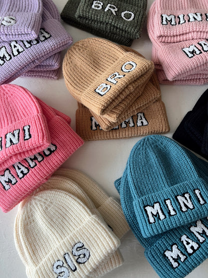 Colorful beanies with text labels like "BRO," "MAMA," "MINI," and "SIS" arranged on a table.