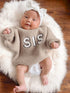 Happy baby wearing a beige sweater with "SIS" text, white bow, lying on a fluffy rug.