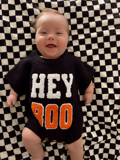 Smiling baby in a black sweater with "HEY BOO" on a checkered blanket.