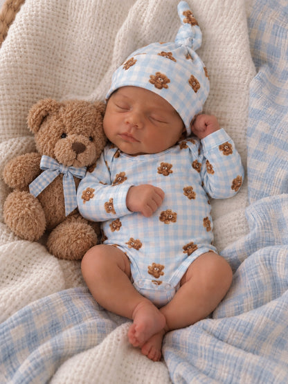 Sleeping baby in a blue bear-patterned outfit and hat, cuddling a teddy bear on a cozy blanket.