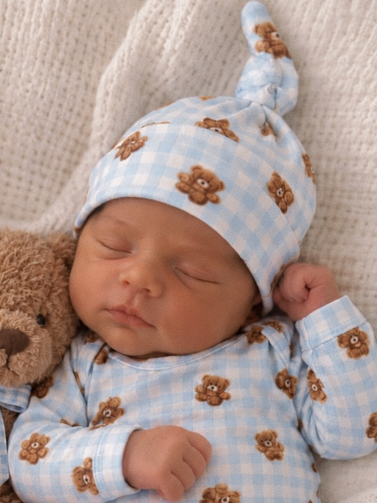 Sleeping newborn in a blue bear-patterned outfit, snuggled with a teddy bear on a cozy blanket.