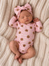 Sleeping baby in pink teddy bear onesie with a large bow, lying on a cozy cream blanket.