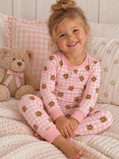 Smiling child in pink pajamas with bear prints, sitting on a cozy blanket next to a teddy bear.