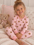 Smiling child in pink pajamas with bear prints, sitting on a cozy blanket next to a teddy bear.