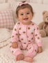 Smiling baby in a pink bear-patterned onesie, sitting on a cozy blanket with a teddy bear in the background.