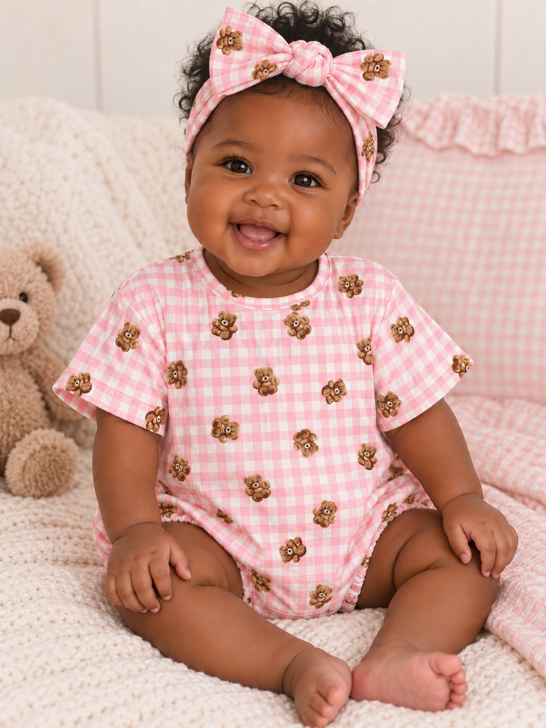 Smiling baby in pink bear-patterned outfit with a matching headband, seated on a cozy blanket near a teddy bear.