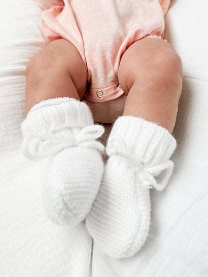 Baby feet in soft white knitted booties, resting on a soft white blanket, wearing a light pink outfit.