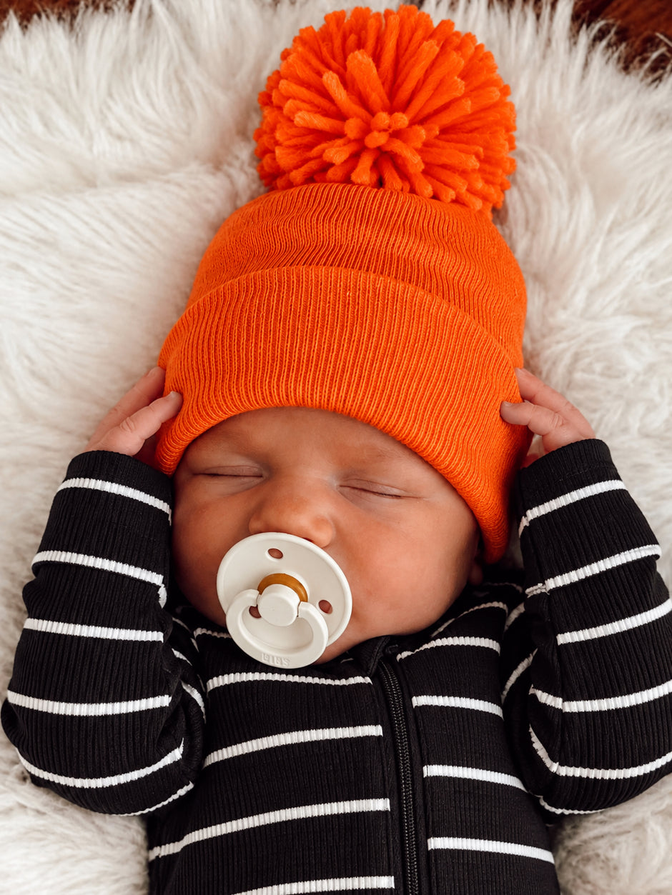 Sleeping baby wearing an orange pom-pom hat and black striped outfit, holding a pacifier on a fluffy blanket.