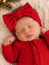Sleeping baby in a red outfit with a large bow, resting on a cozy blanket with festive decorations.