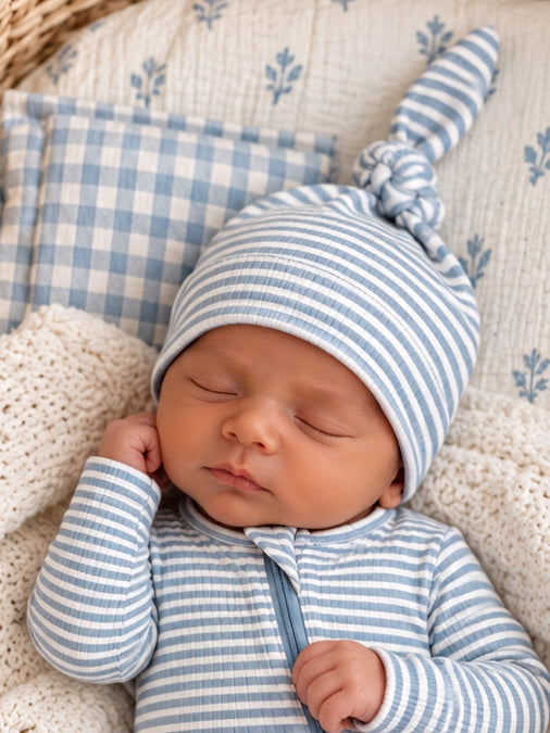 Sleeping newborn in blue striped outfit and knotted hat, resting on a cozy blanket and patterned pillows.