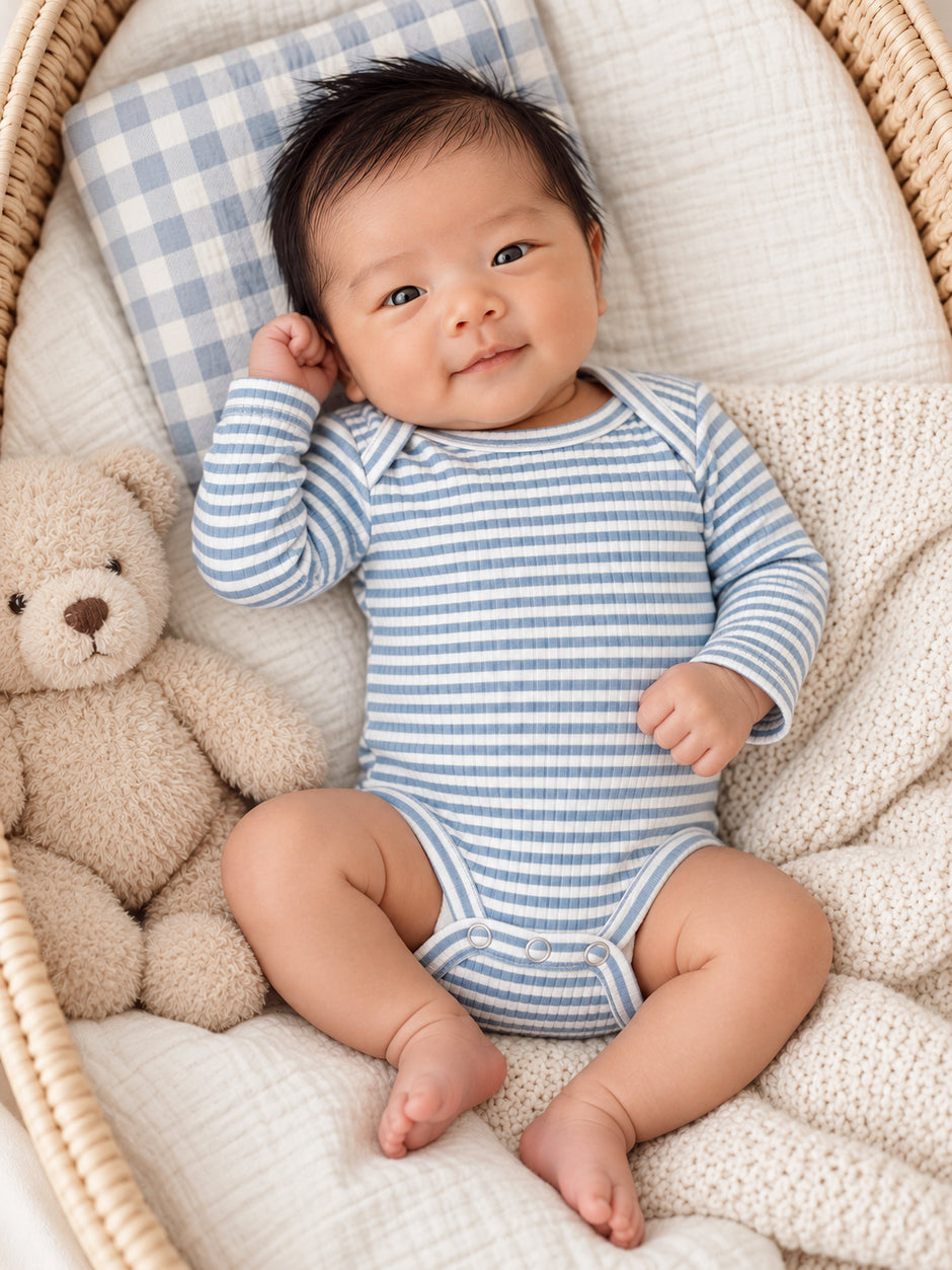 Smiling baby in a striped onesie, resting in a basket with a teddy bear and a checkered pillow.