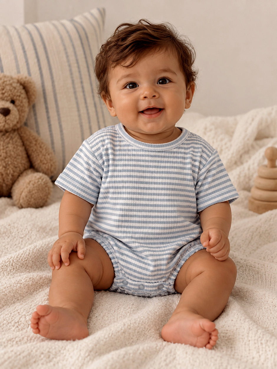 Smiling baby in a striped onesie sitting on a cozy blanket with a teddy bear nearby.